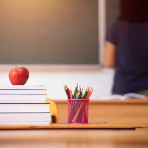 Pencils, apple and books, teacher and blackboard in the background.