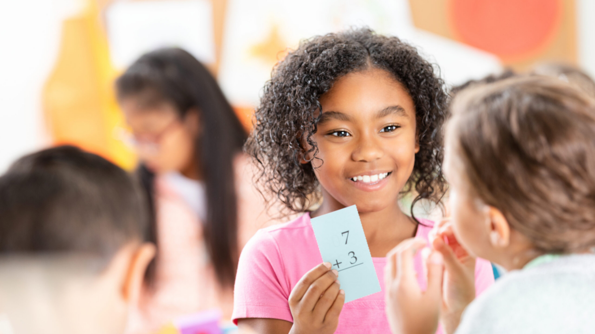 Children engaged in an indoor classroom activity. A girl holds a math card, smiling at her peer, with others learning.