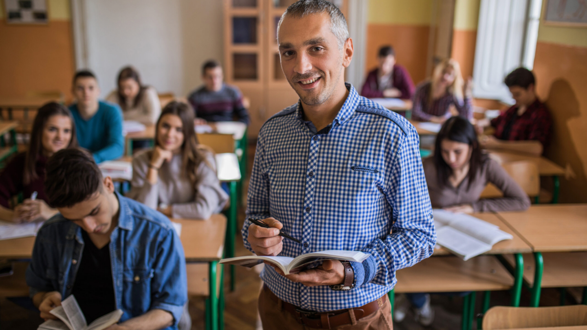 Smiling professor standing in the classroom and looking at camera while students are learning in the background.